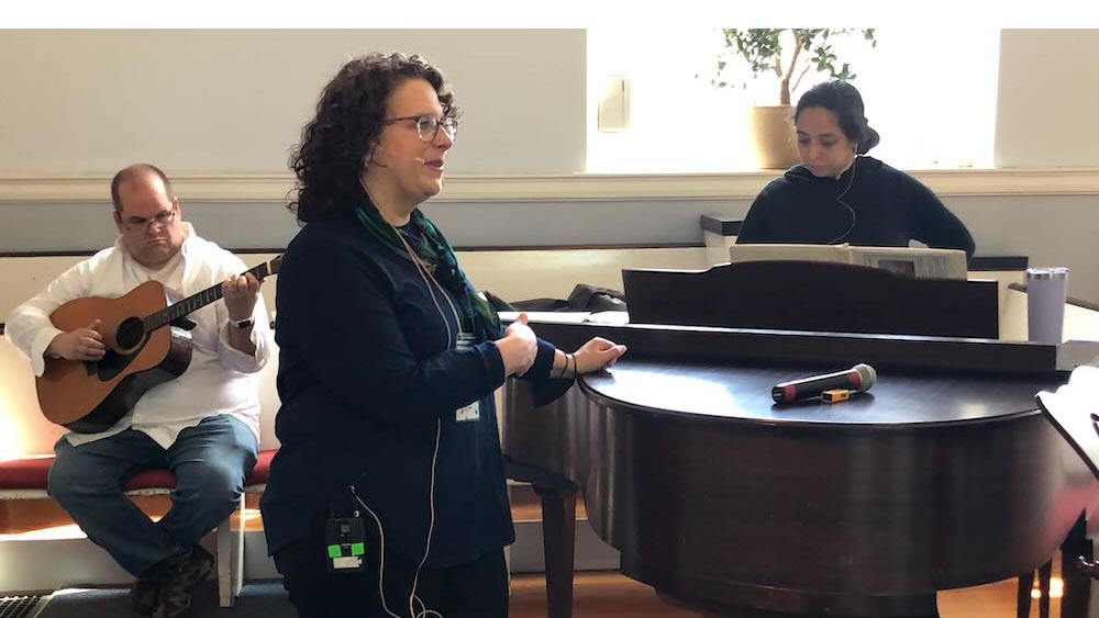 Man playing guitar, woman playing piano, and woman standing by piano facing congregation