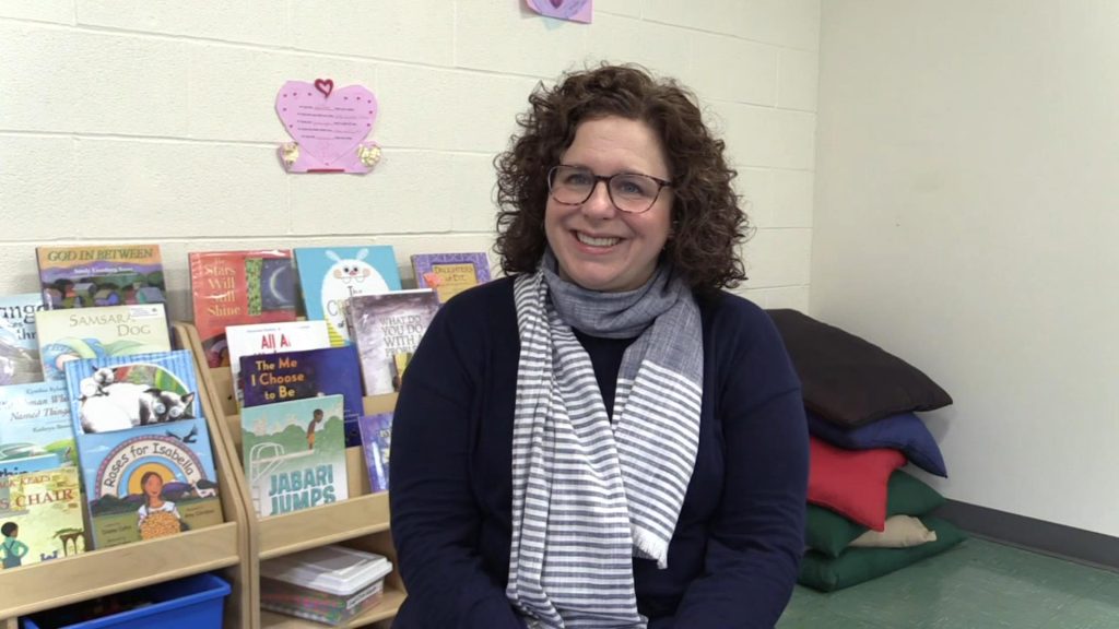 Elizabeth Baldes, smiling adult white woman with black curly hair, glasses, and a scarf seated in front of children's books. 