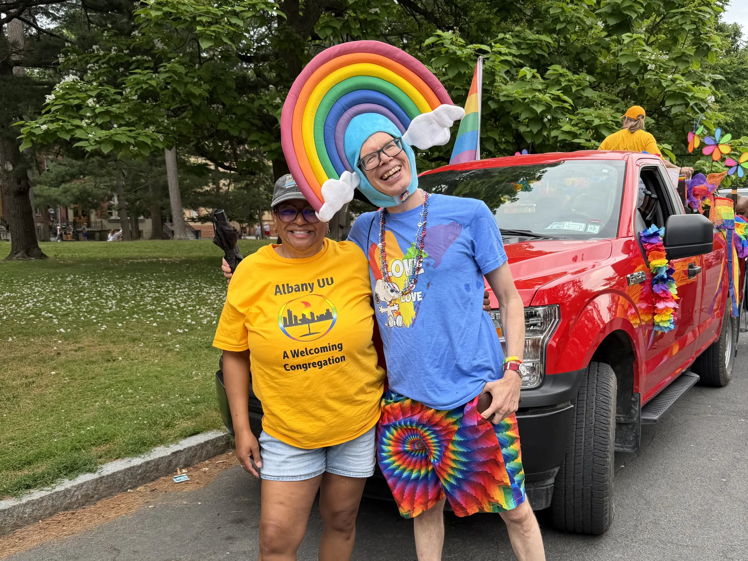 Two people posing in front of a decorated red pickup truck with a rainbow wing headpiece at a Pride parade