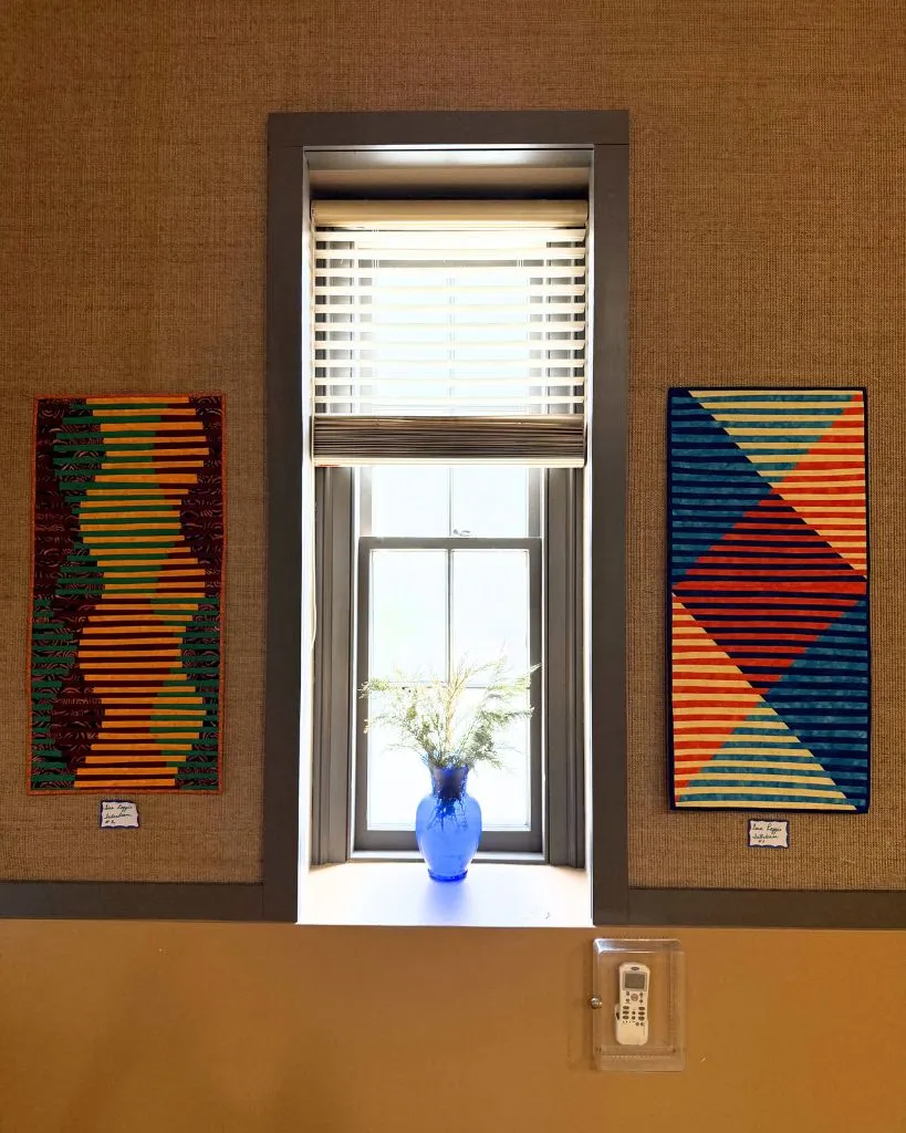 Quilted fabric art panels flanking a window with a blue glass vase of greenery on the sill