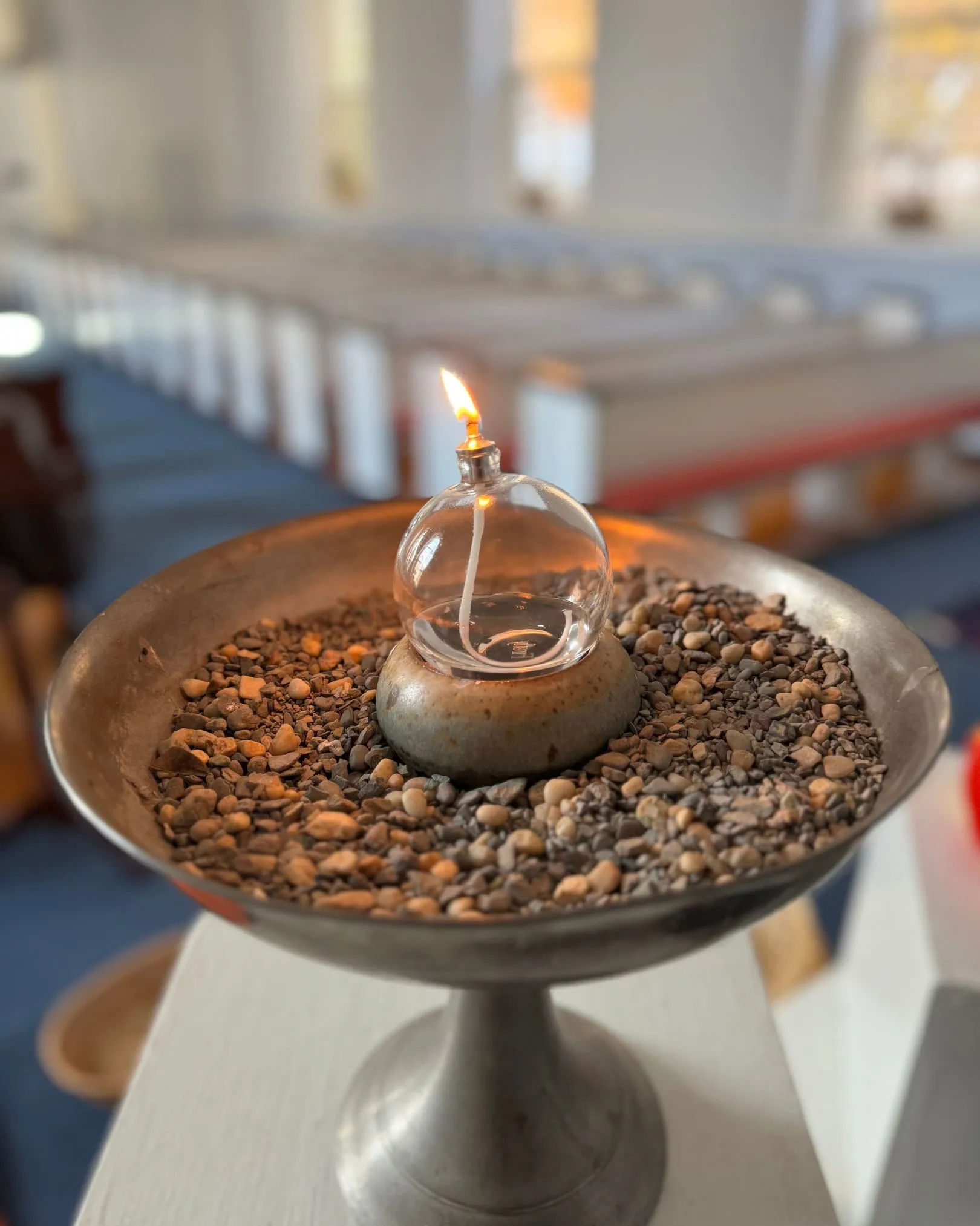 A lit oil candle in a glass globe set among stones in a bronze bowl, with sanctuary pews in the background