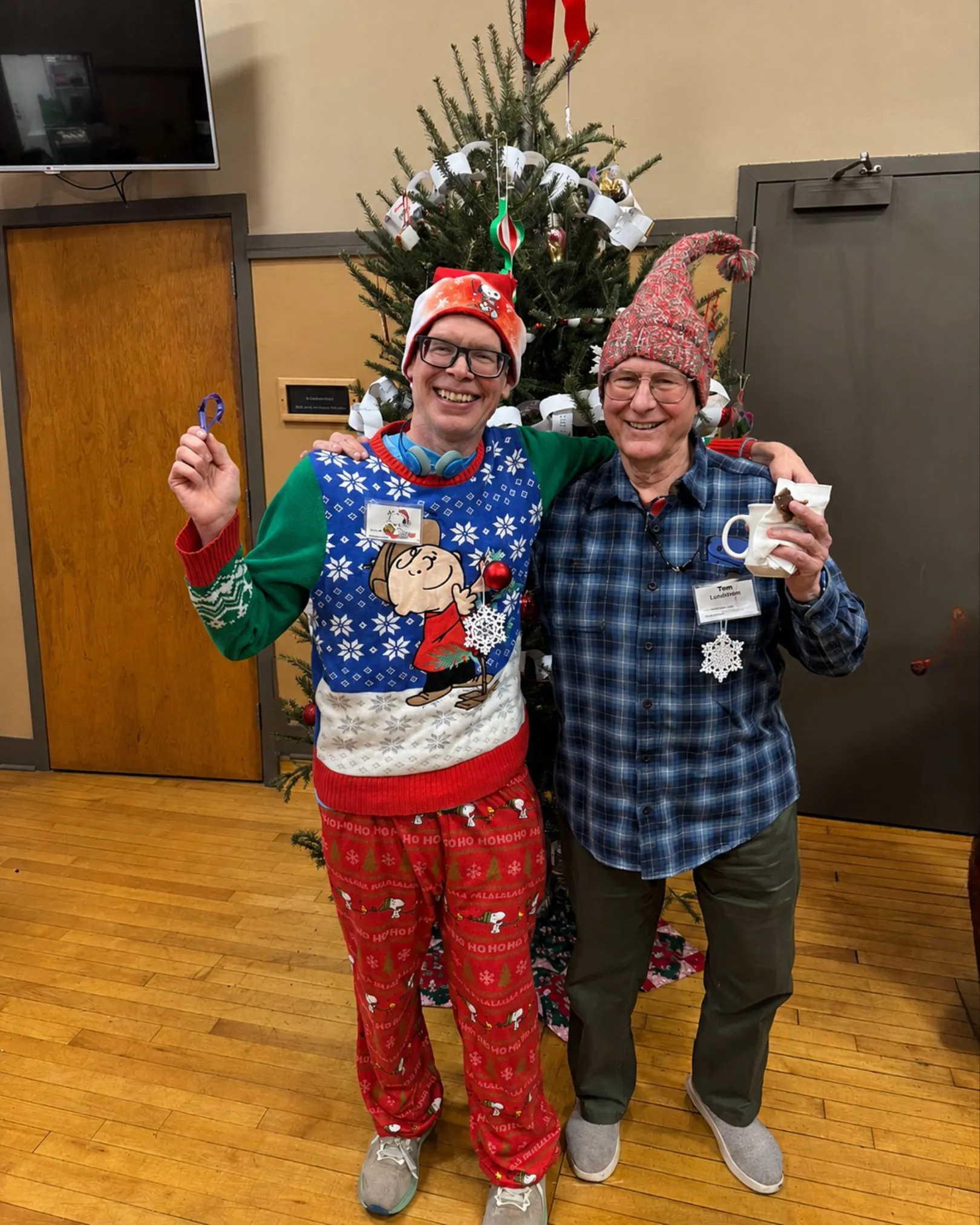 Two men in Christmas-themed sweaters and hats posing in front of a decorated Christmas tree