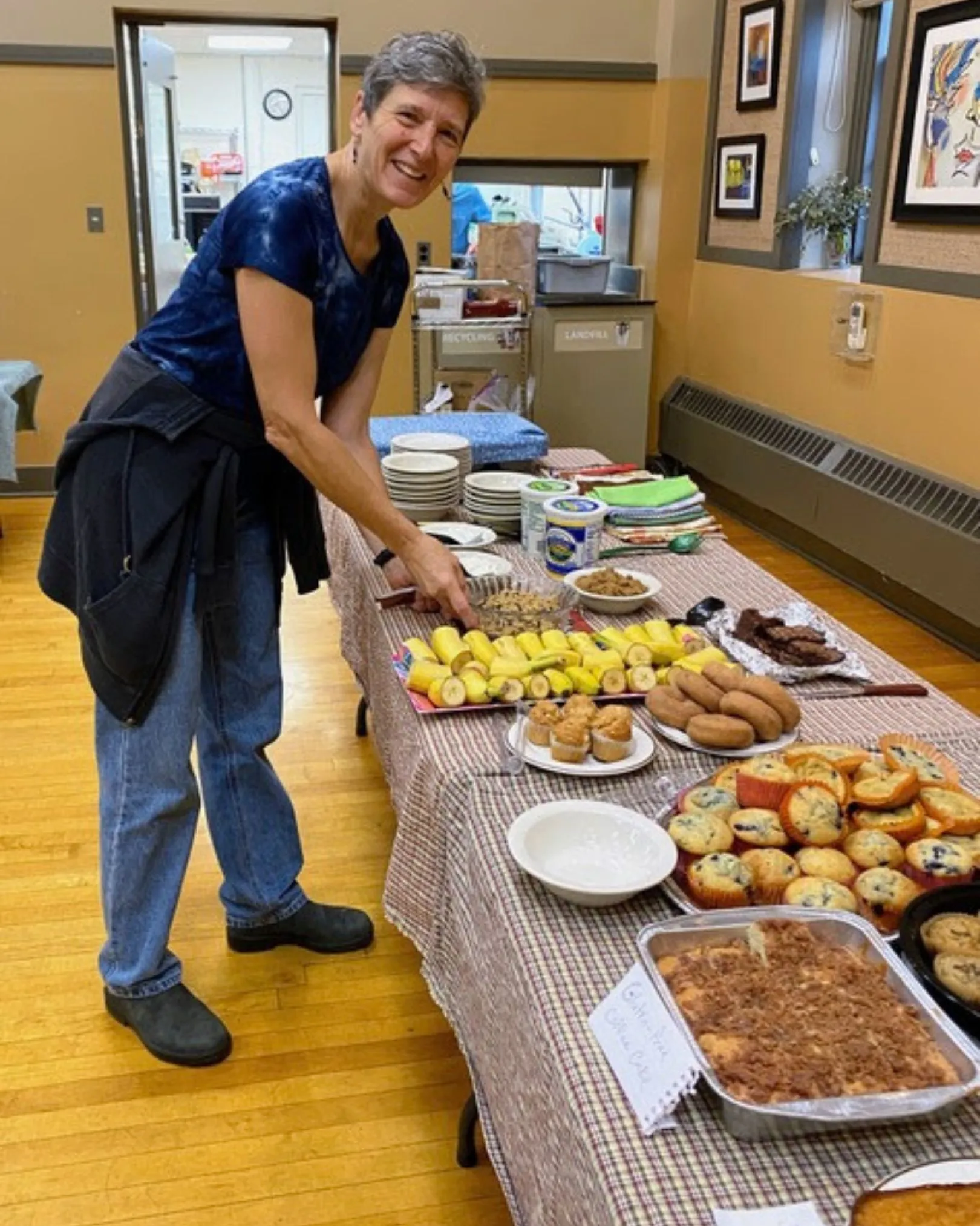 A volunteer serving herself from a breakfast buffet of bananas, muffins, and pastries