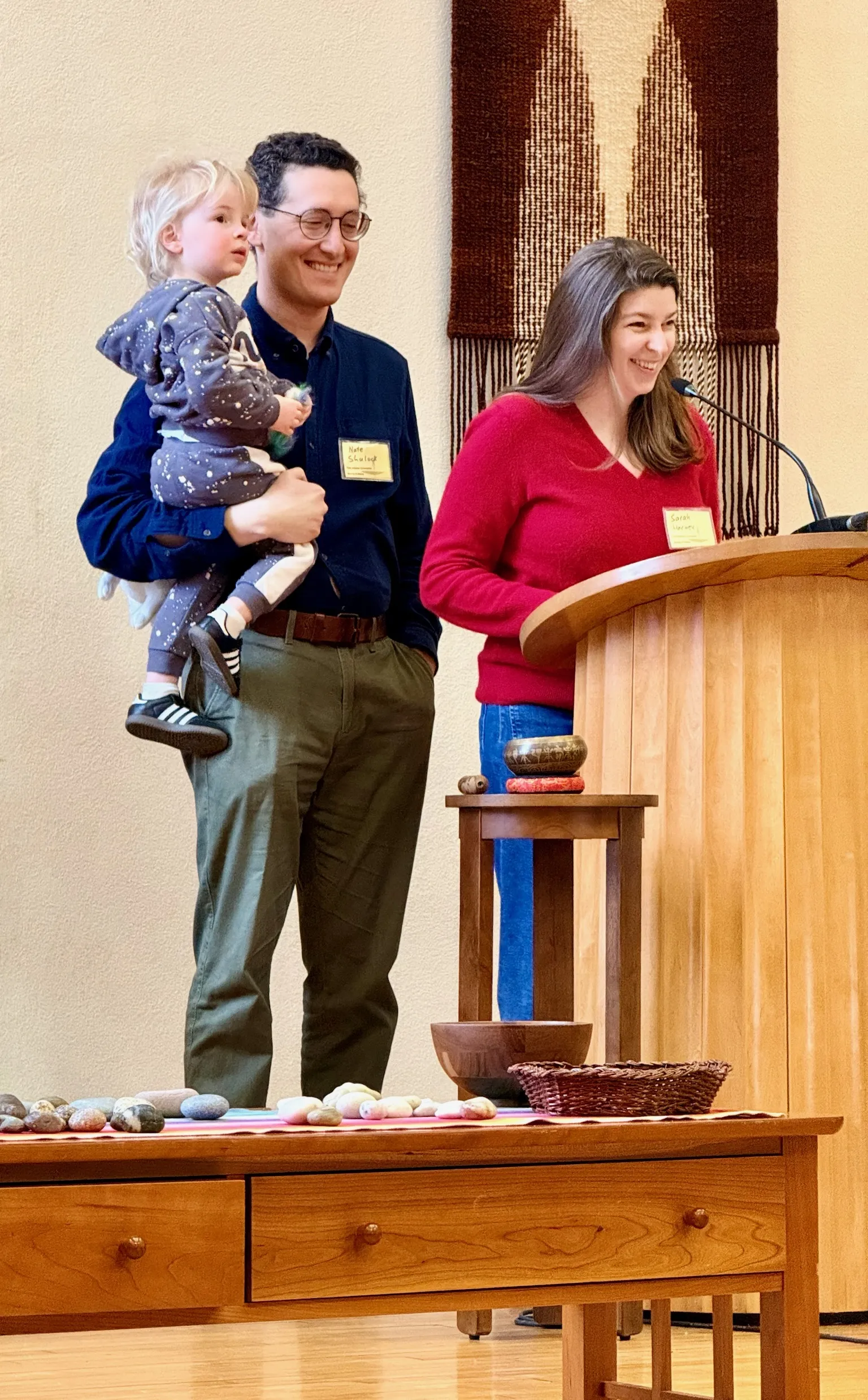 A father holding a toddler beside a woman speaking at a podium during a service