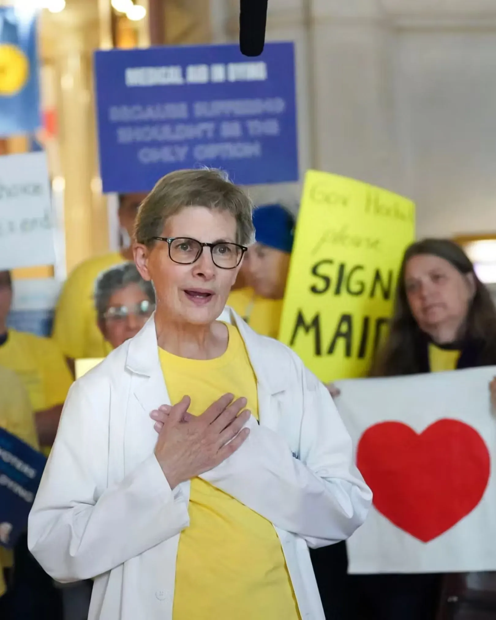 A speaker in a white coat with her hand over her heart addressing a rally with a 'Sign MAID' sign behind her