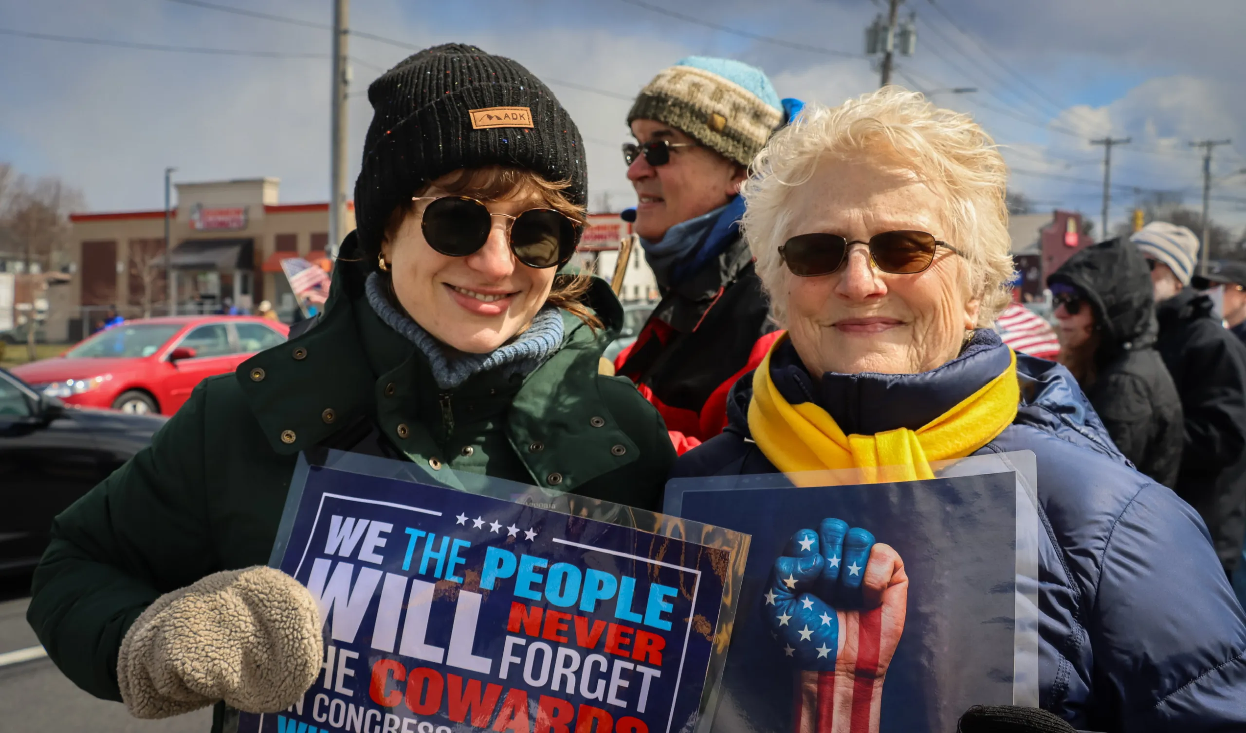 Two women holding 'We The People Will Never Forget' and 'Ejecting Kings Since 1776' signs at a protest