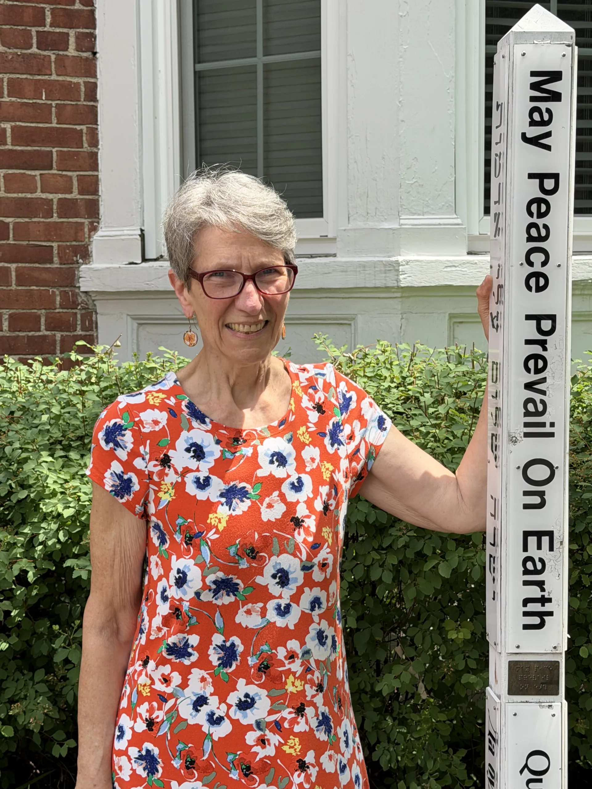 Paula Brewer smiling beside a 'May Peace Prevail on Earth' peace pole