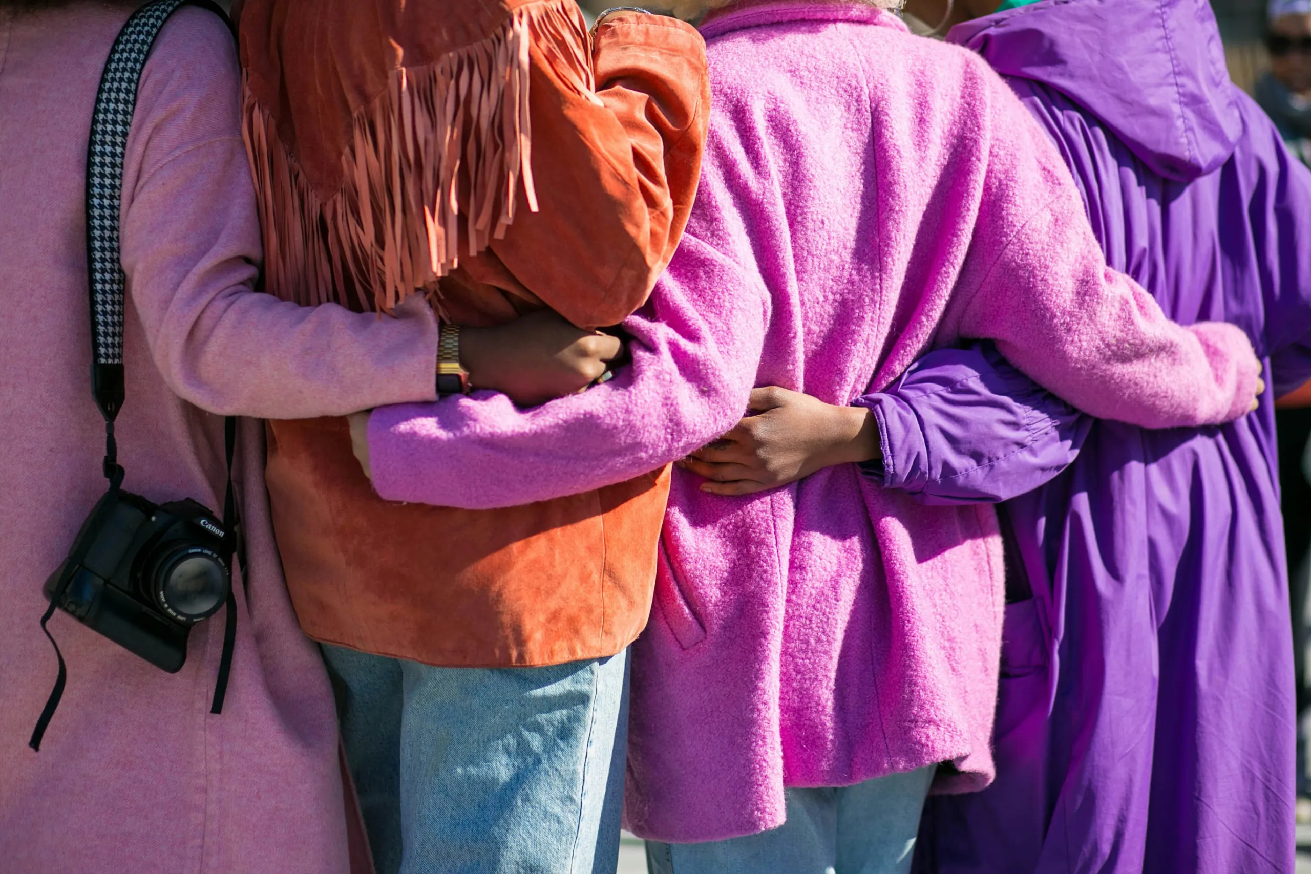 A row of people in pink and purple coats with arms around each other's backs, seen from behind