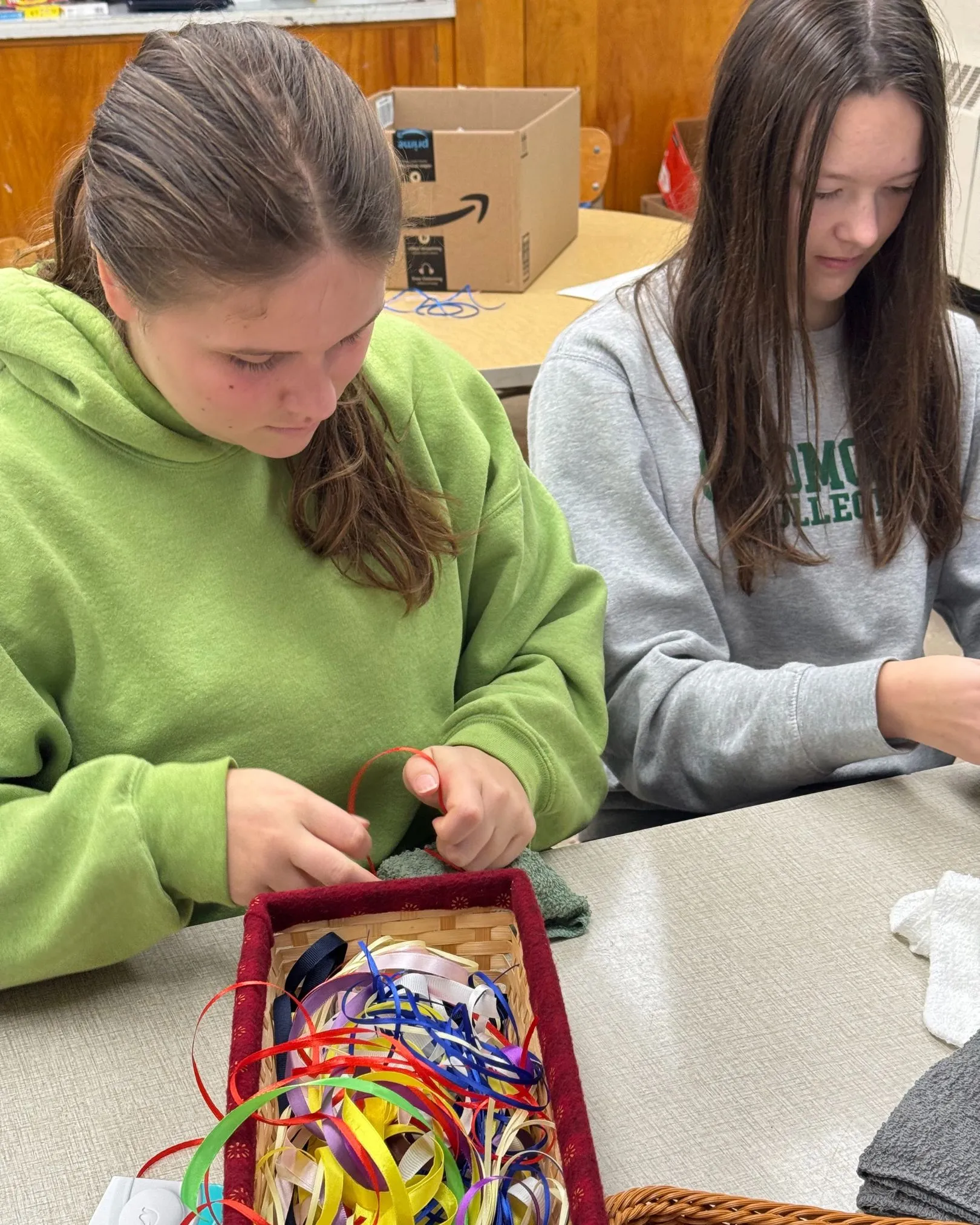 Two girls tying colorful ribbons at a table with a basket of craft ribbon