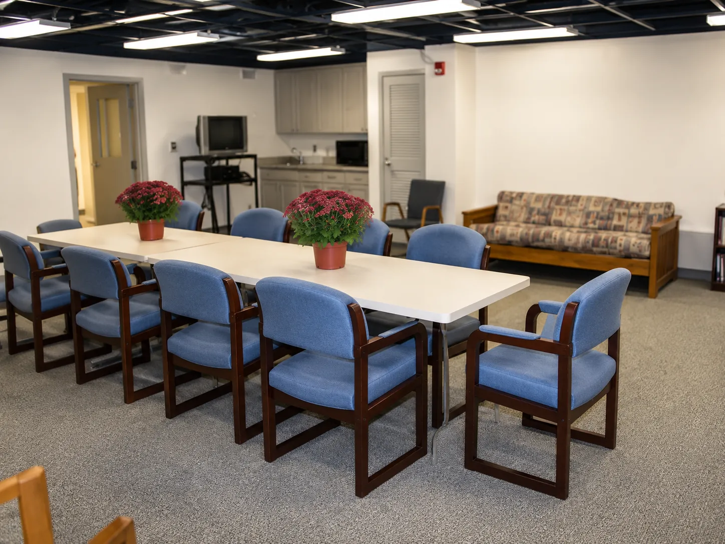 A long table with blue upholstered chairs, potted mums, and a kitchenette in a classroom