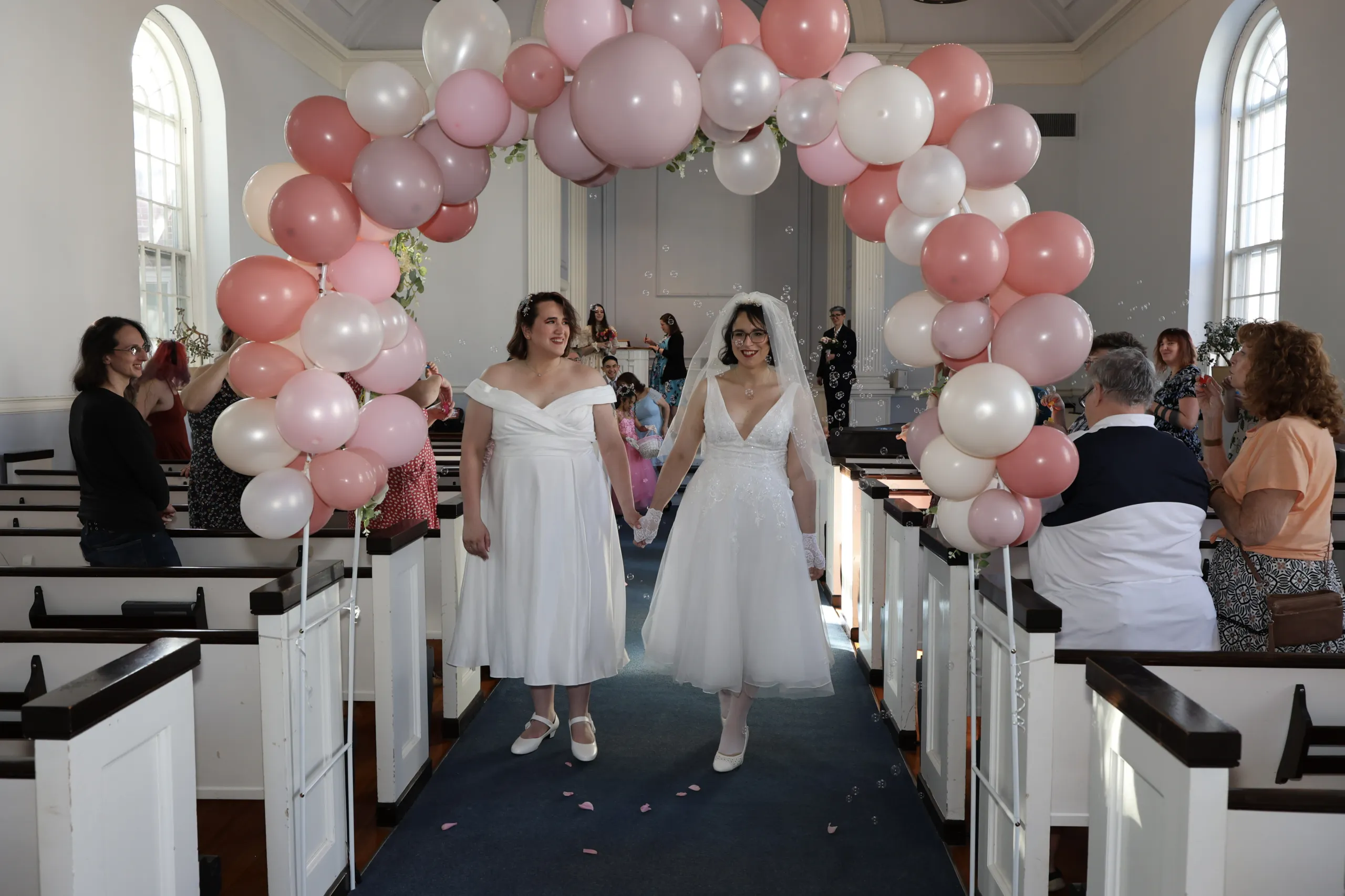 Two brides holding hands and walking together down the aisle under a pink and white balloon arch