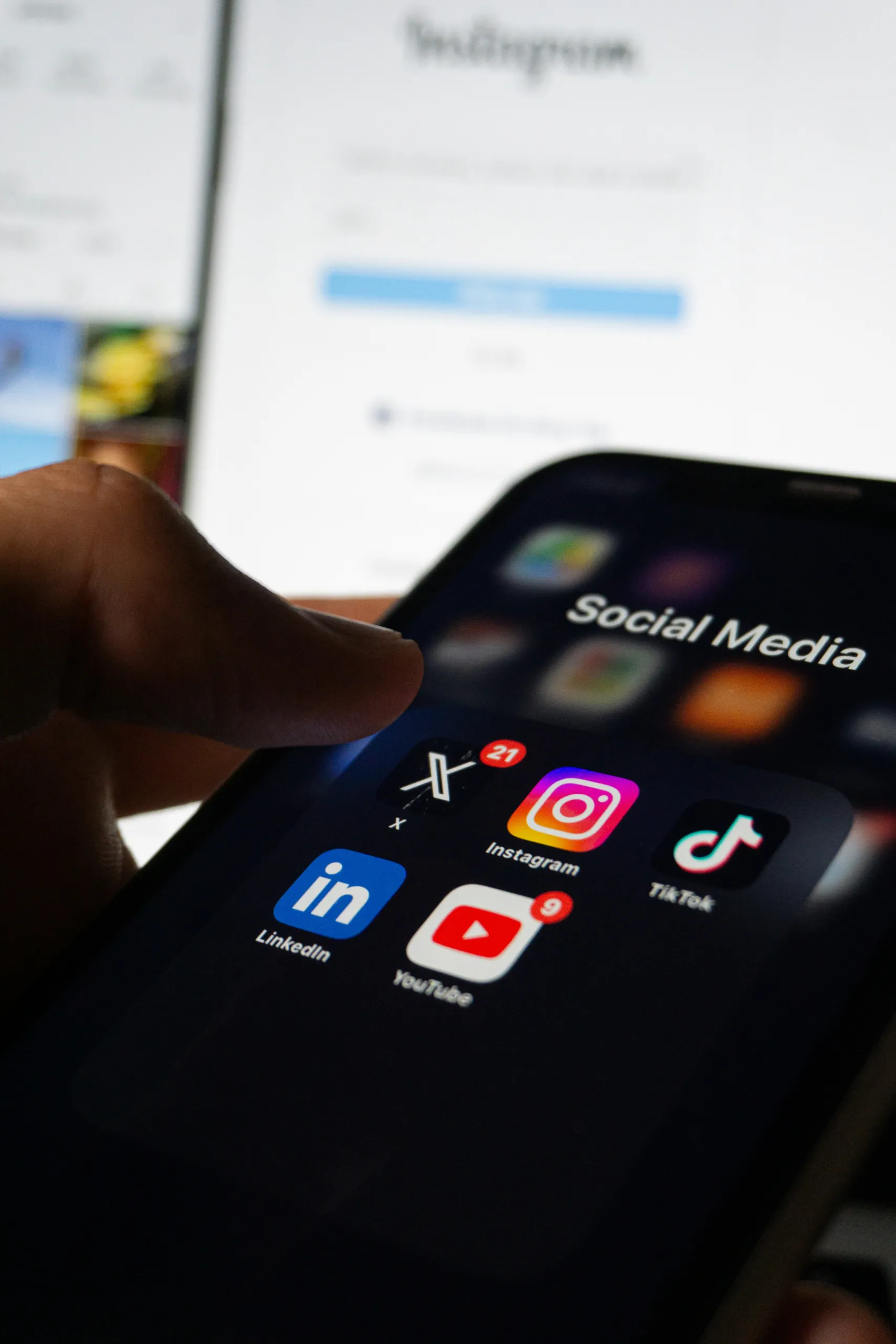 A hand holding a phone displaying social media app icons in front of a laptop screen