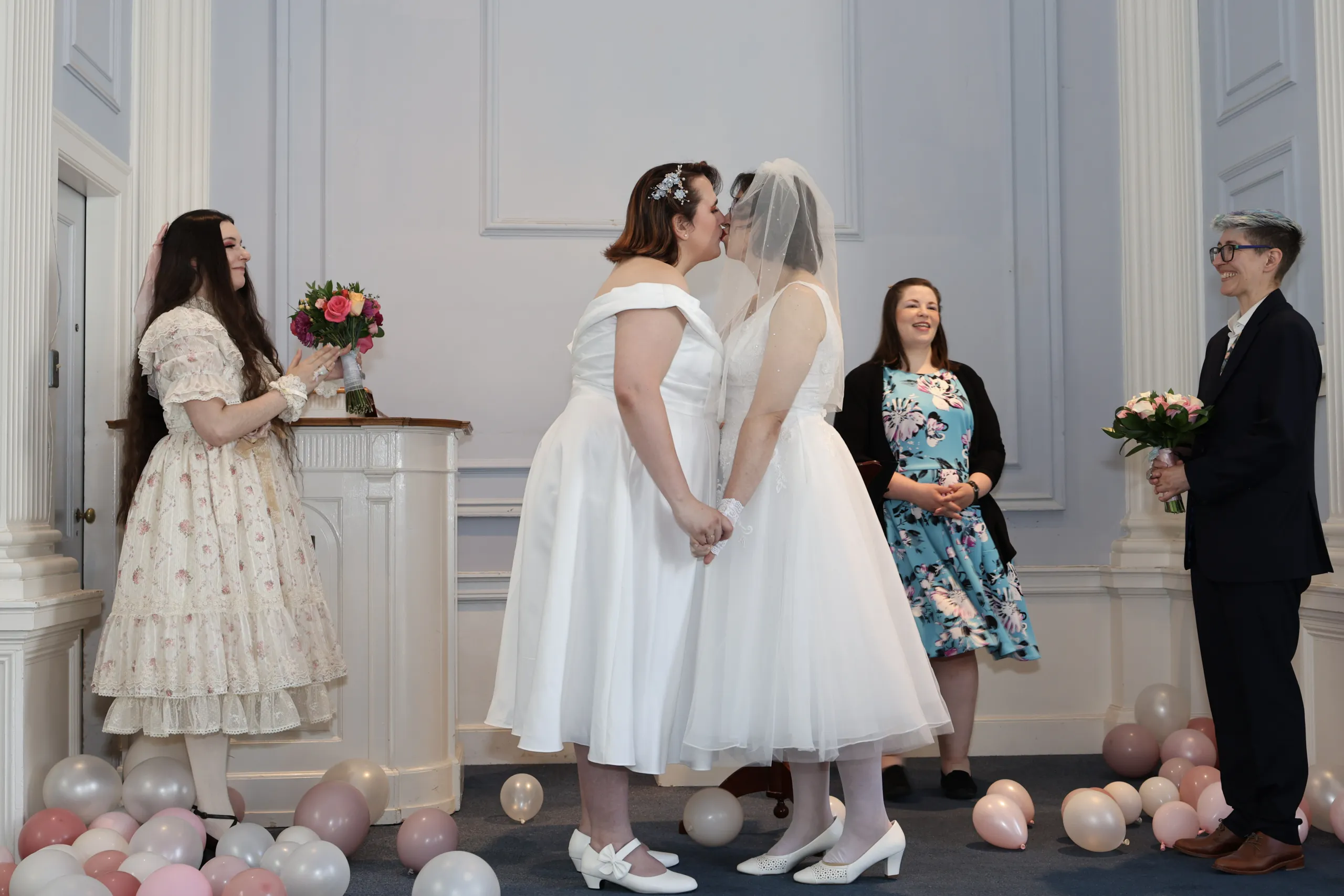 Two brides kissing at the altar as an officiant and attendant look on, surrounded by balloons