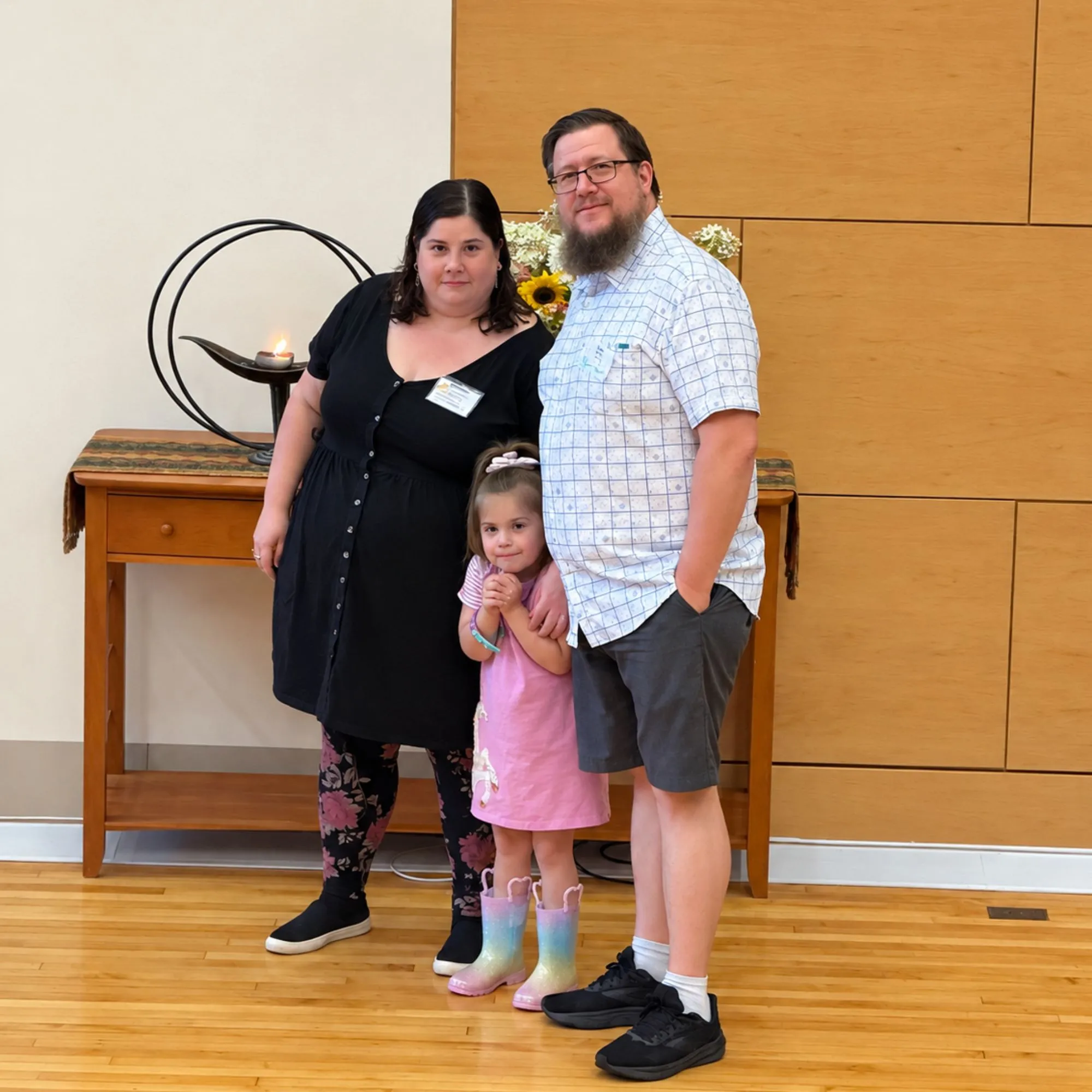 A family of three standing beside a lit chalice and flowers