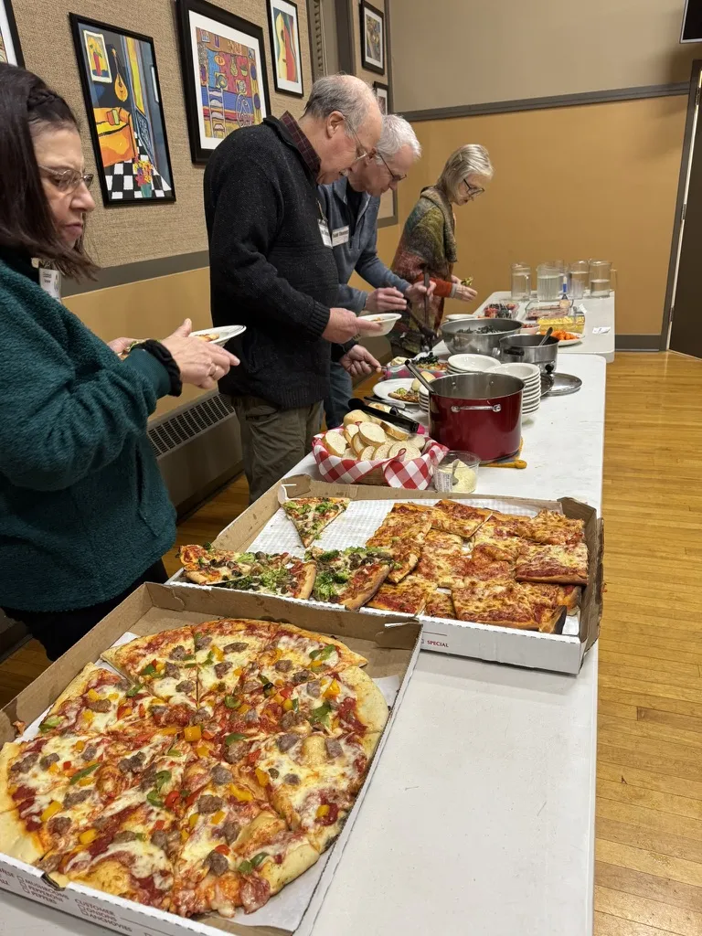 People serving themselves pizza and potluck dishes from a buffet table at a church potluck