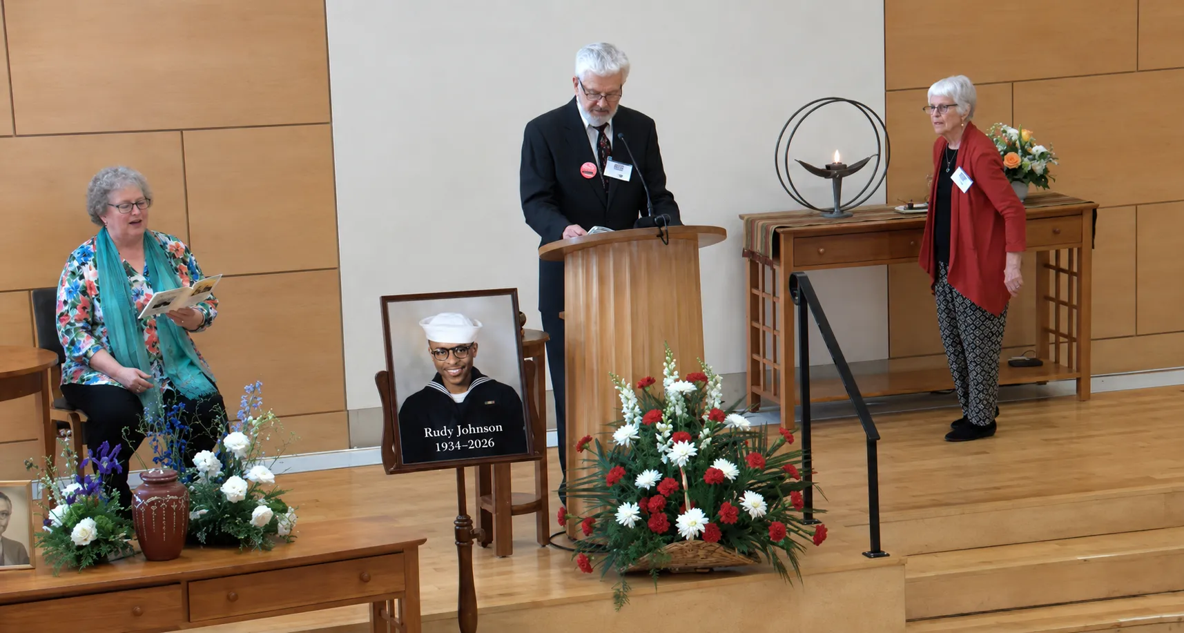 A memorial service program with a photo of Rudy Johnson displayed on an easel beside a speaker at a podium