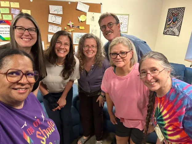 Seven smiling members of the ministerial search committee posing together in a room with a bulletin board and children's artwork