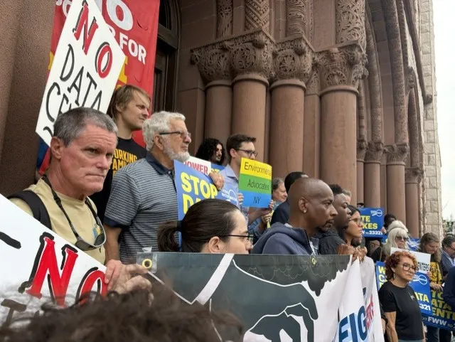 Protesters holding "No Data Center" and "Stop Data" signs outside a stone building