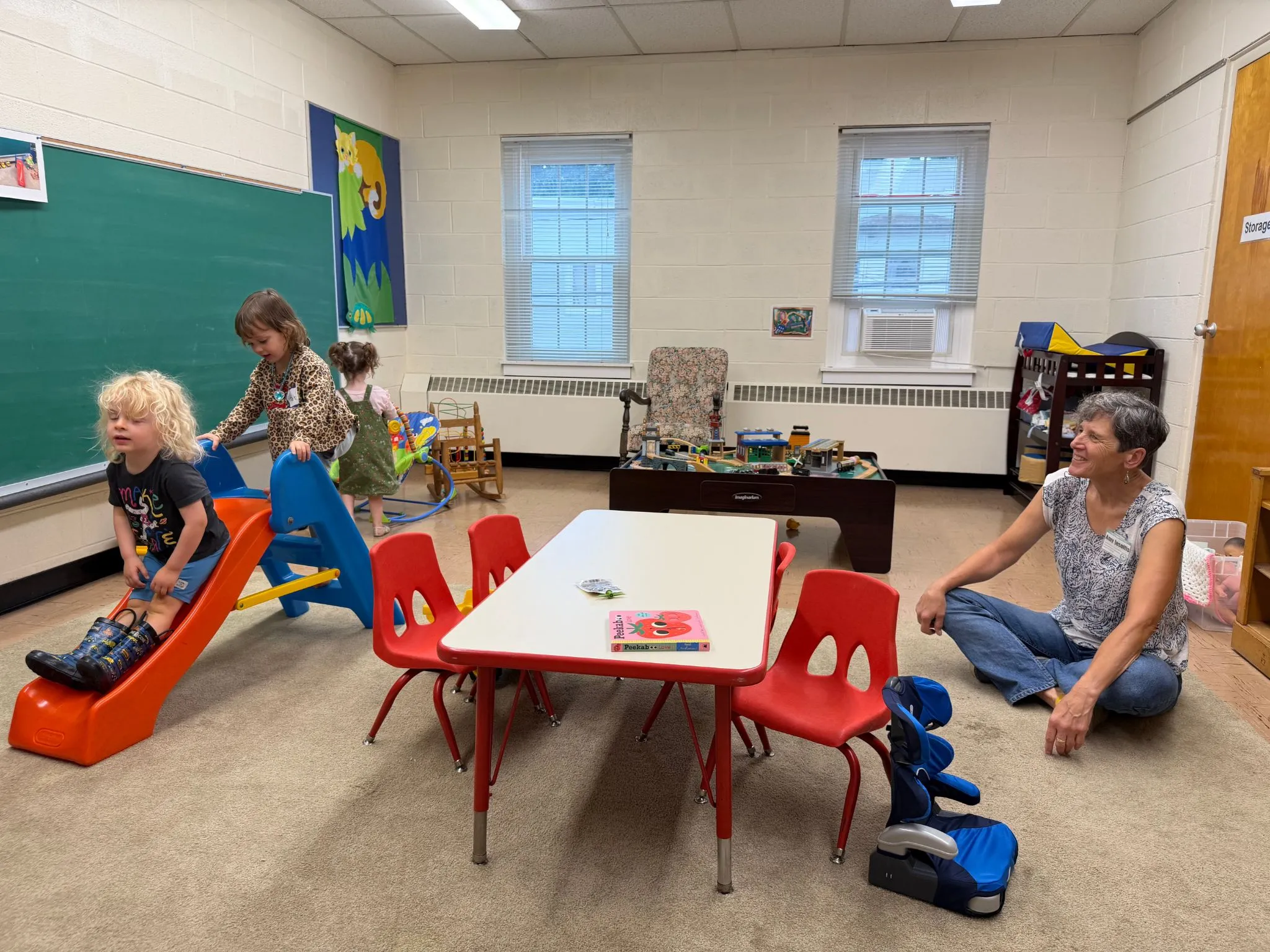 Children playing with a slide and toys in a nursery room while a caregiver sits nearby