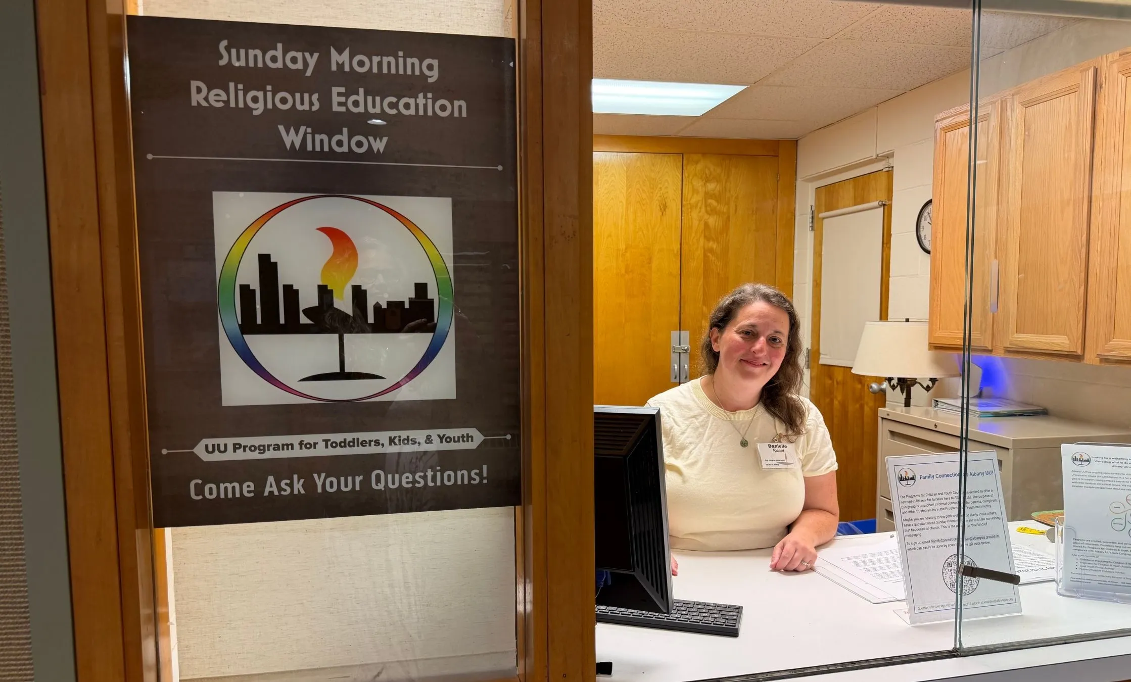 A religious education volunteer smiling at a welcome window decorated with a rainbow flaming chalice sign