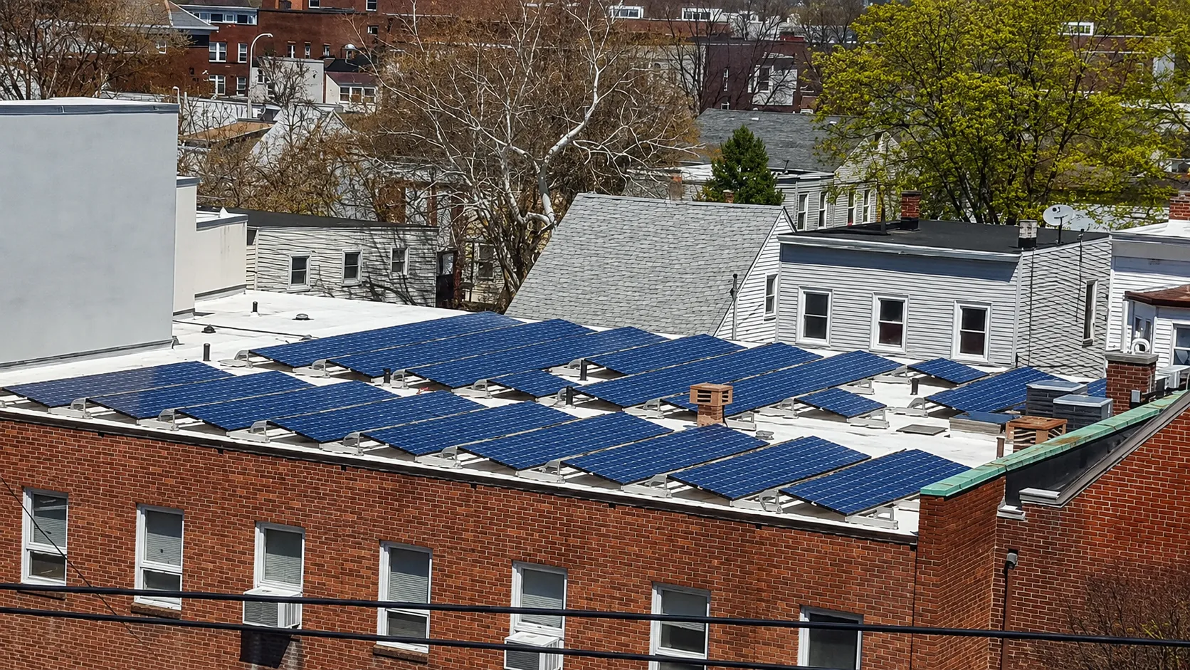 Rows of solar panels on the flat roof of a brick building in a residential neighborhood
