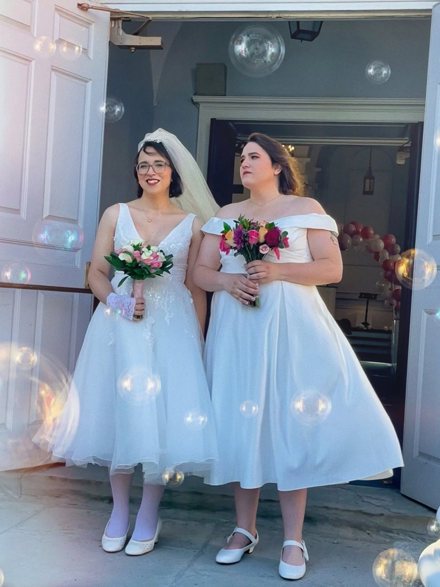 Two brides in white dresses holding bouquets outside a doorway decorated with bubbles