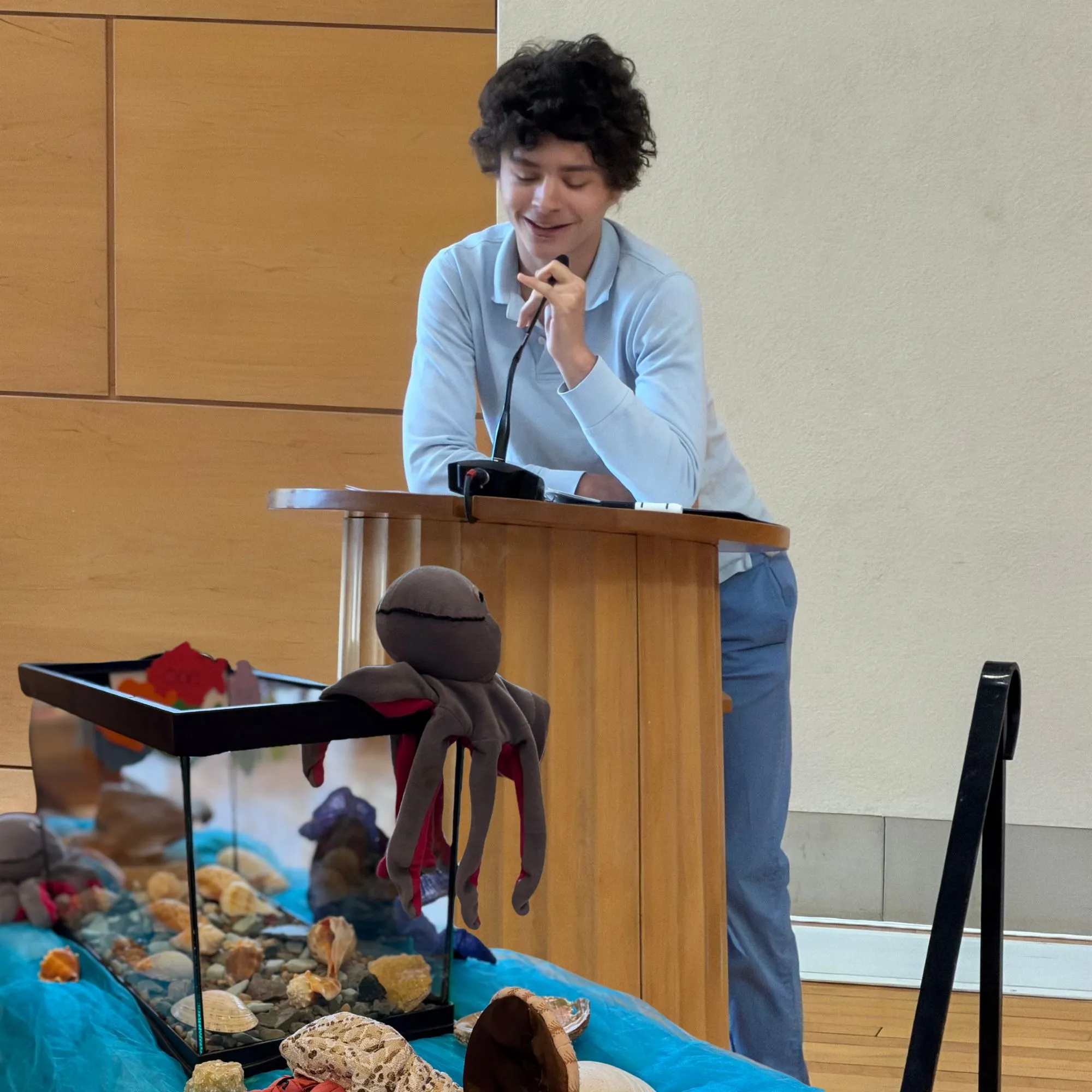 A young person speaking at a podium beside a fish tank decorated with a stuffed octopus toy
