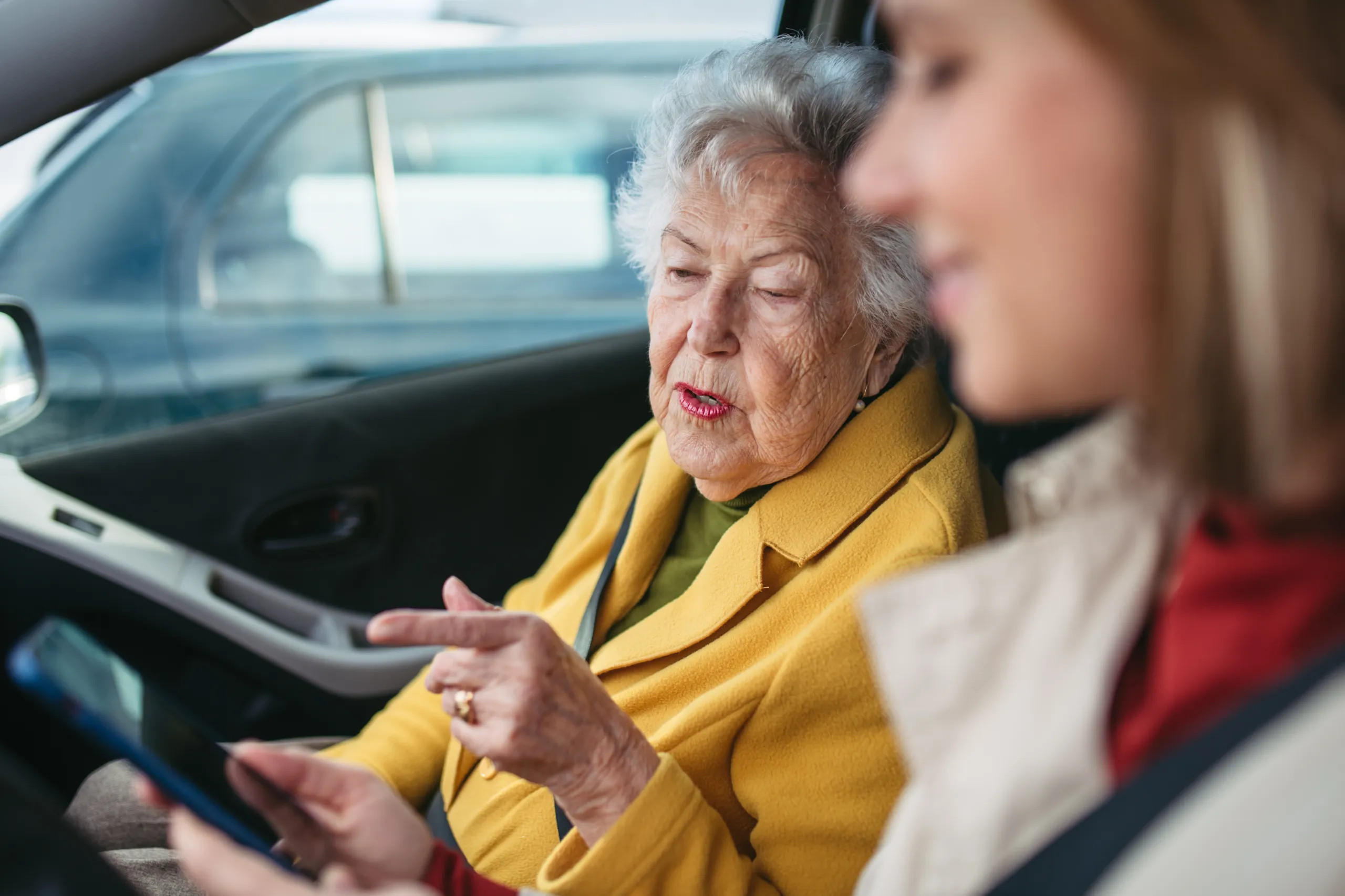 An older woman in a yellow coat pointing at a phone screen while a volunteer driver sits beside her in a car