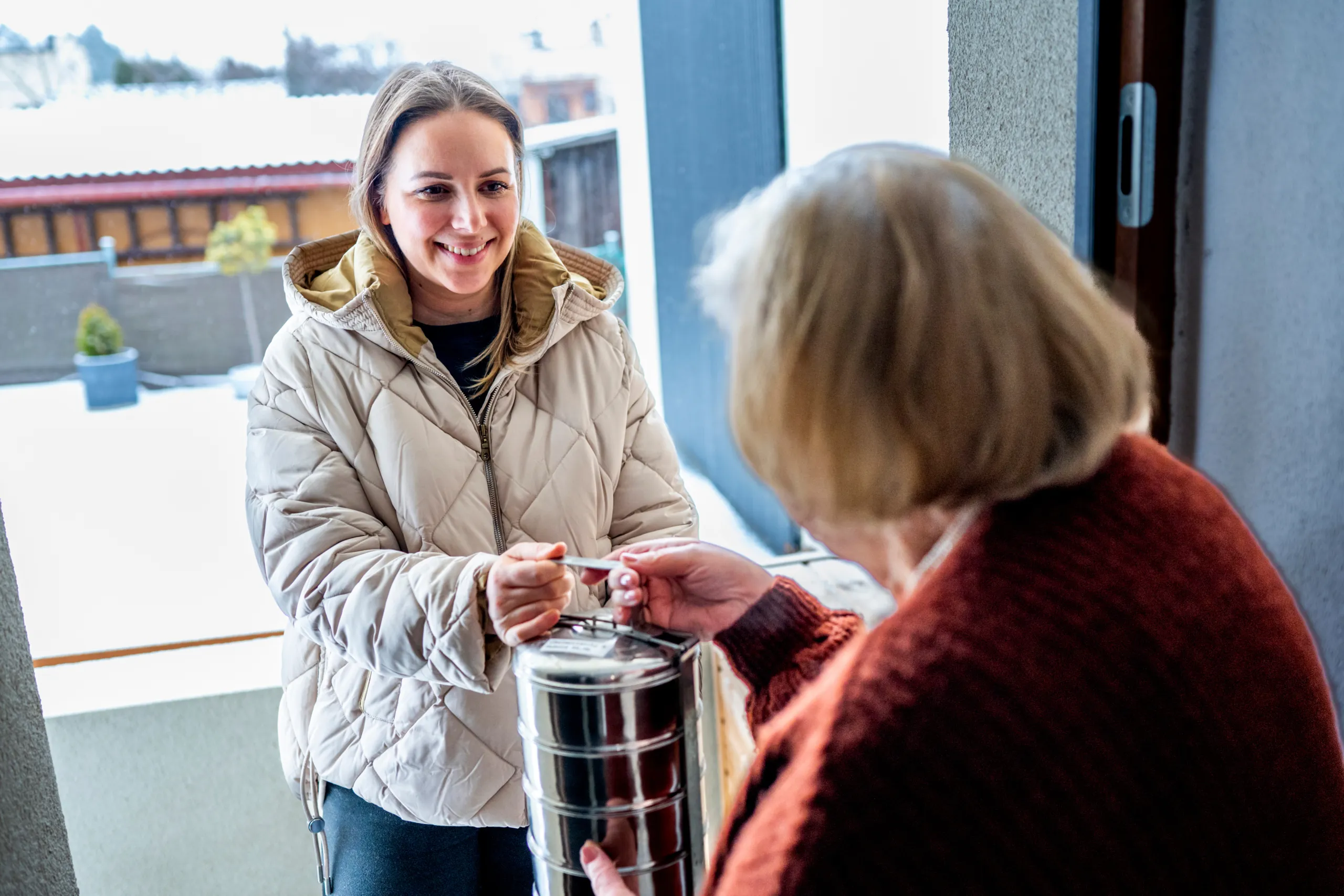 A smiling woman in a quilted jacket handing a stacked metal food carrier to an older woman at her doorway