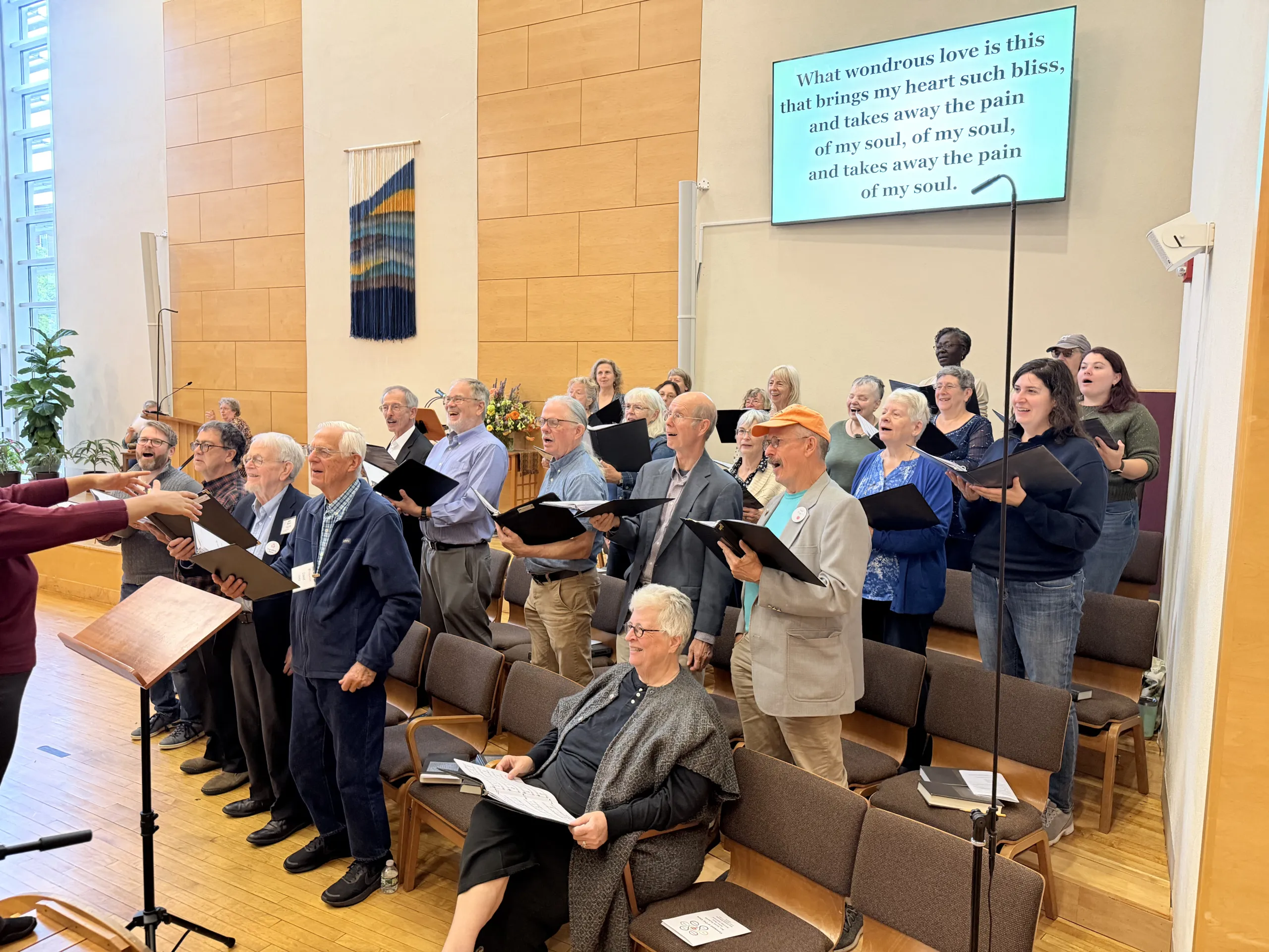 The choir standing and singing from black folders in the sanctuary while the conductor's hands lead from the left