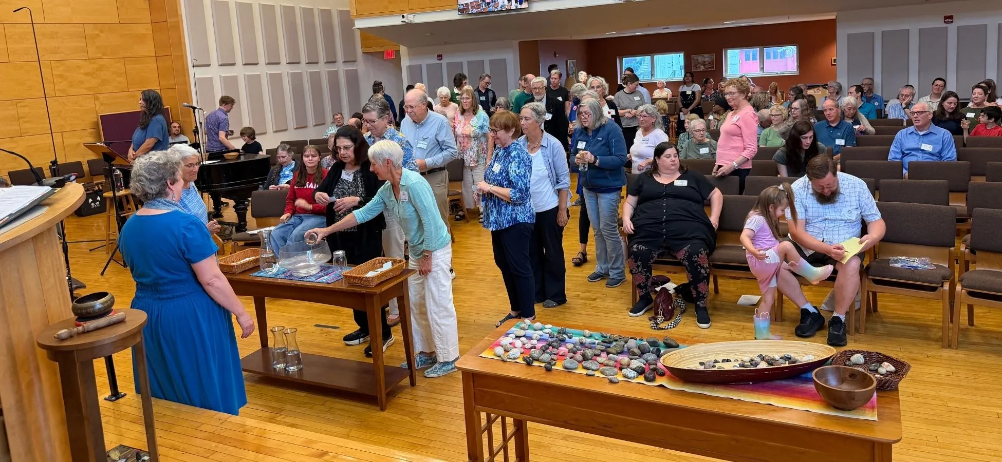 Congregation members lining up to pour water into a glass bowl during a water communion service, with a table of stones in the foreground