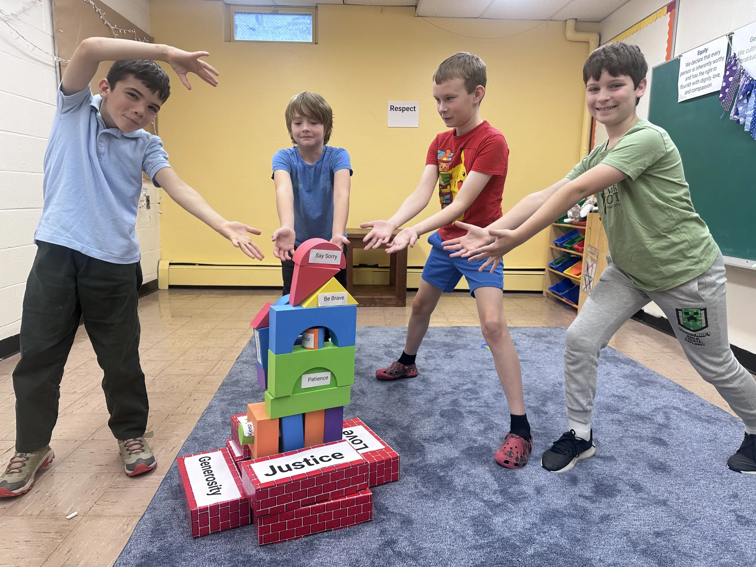 Four boys reaching their hands toward a tower of colorful foam blocks labeled with values like Justice, Generosity, Love, Patience, and Be Brave