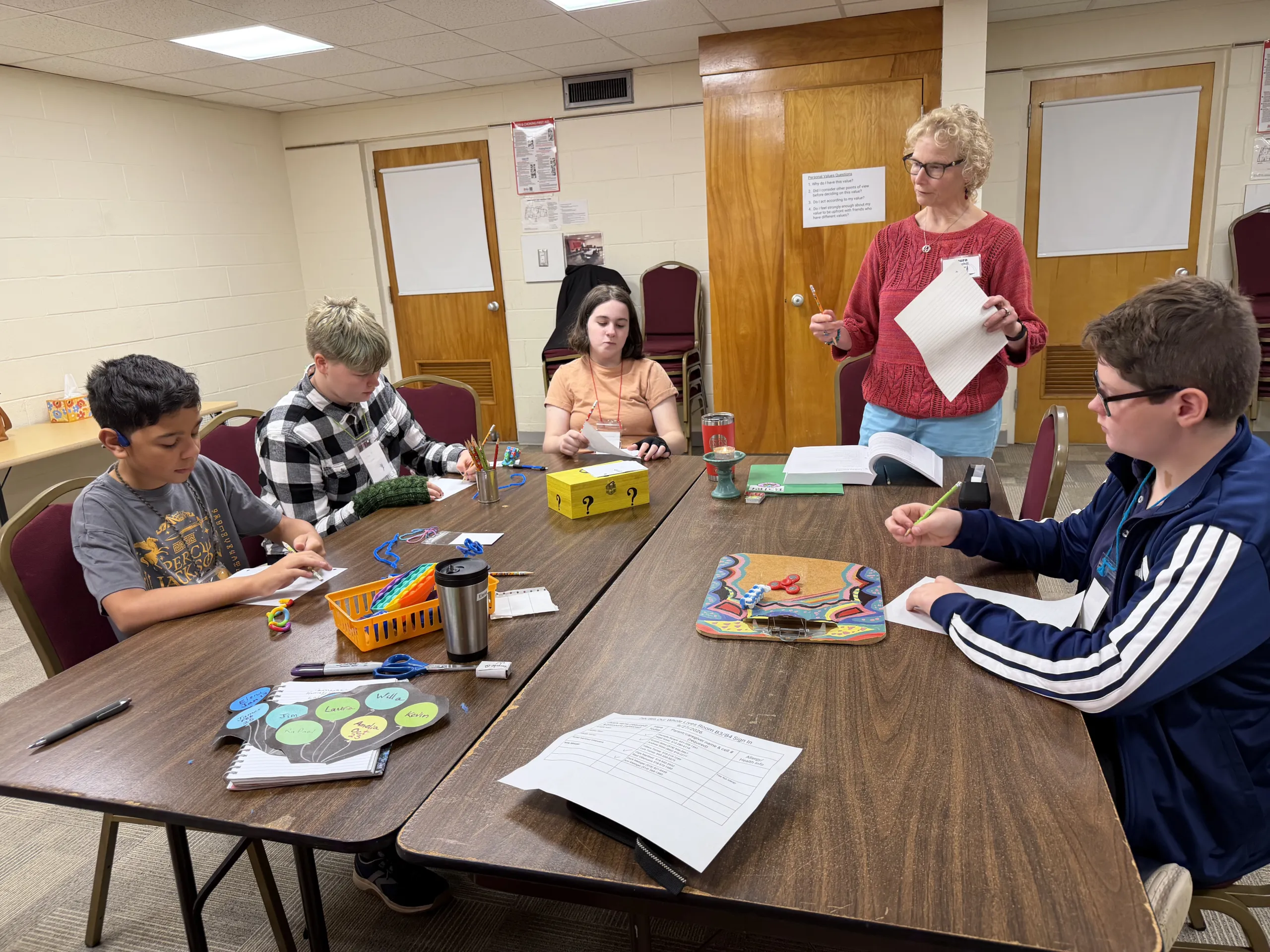 Four middle school youth drawing and writing at a table while an adult volunteer stands and reads from a page